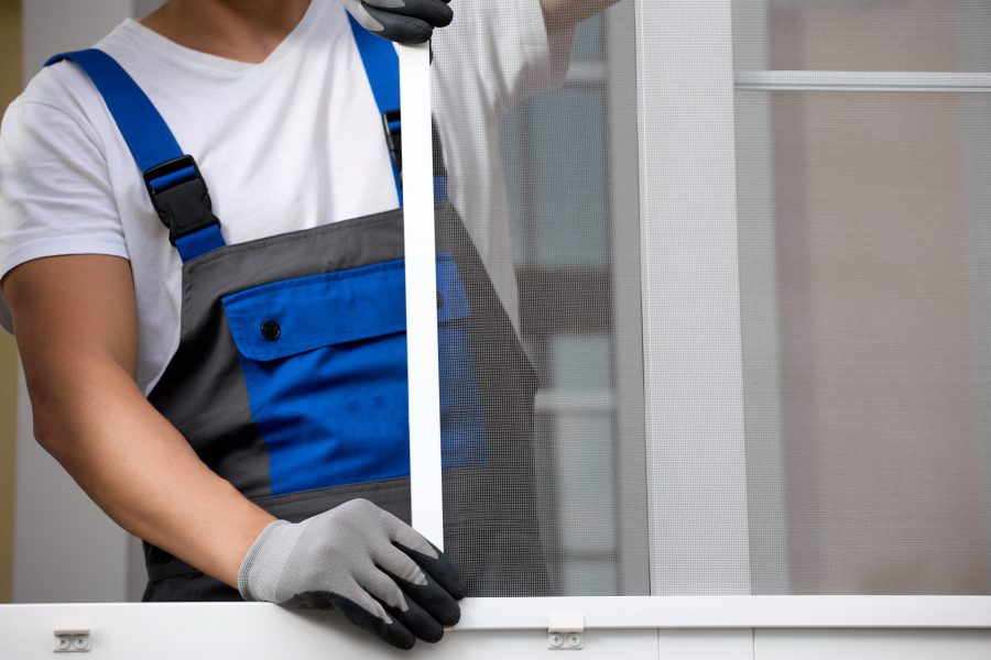 Advertising professional installation of window systems and mosquito nets. Man in a work jumpsuit and a white T-shirt with gloves on his hand installs a protective insect net on a plastic window